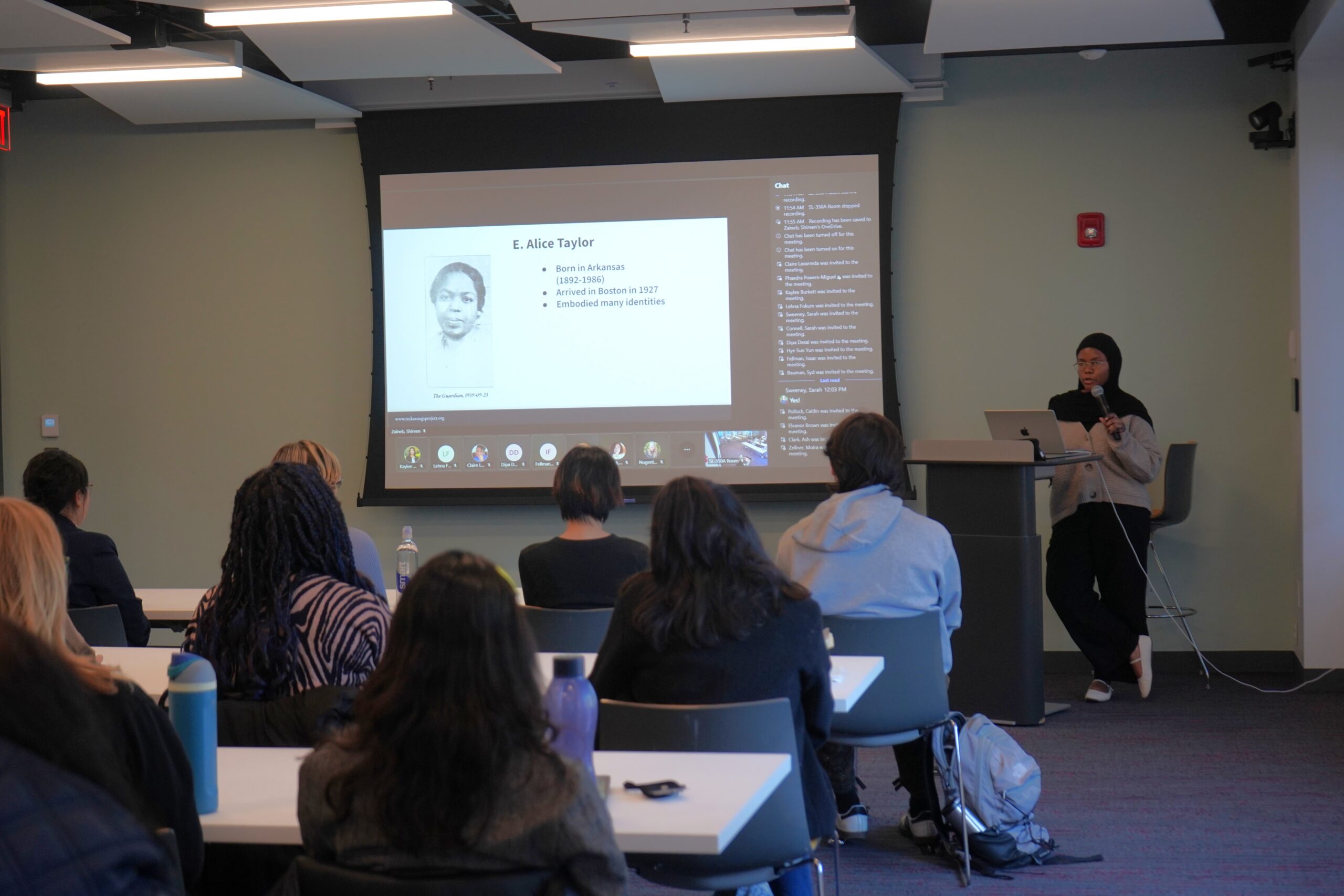 Safia discussing her research on E. Alice Taylor. A person standing by a podium holding a microphone in front of projected slides. There are nine audience members visible from behind.