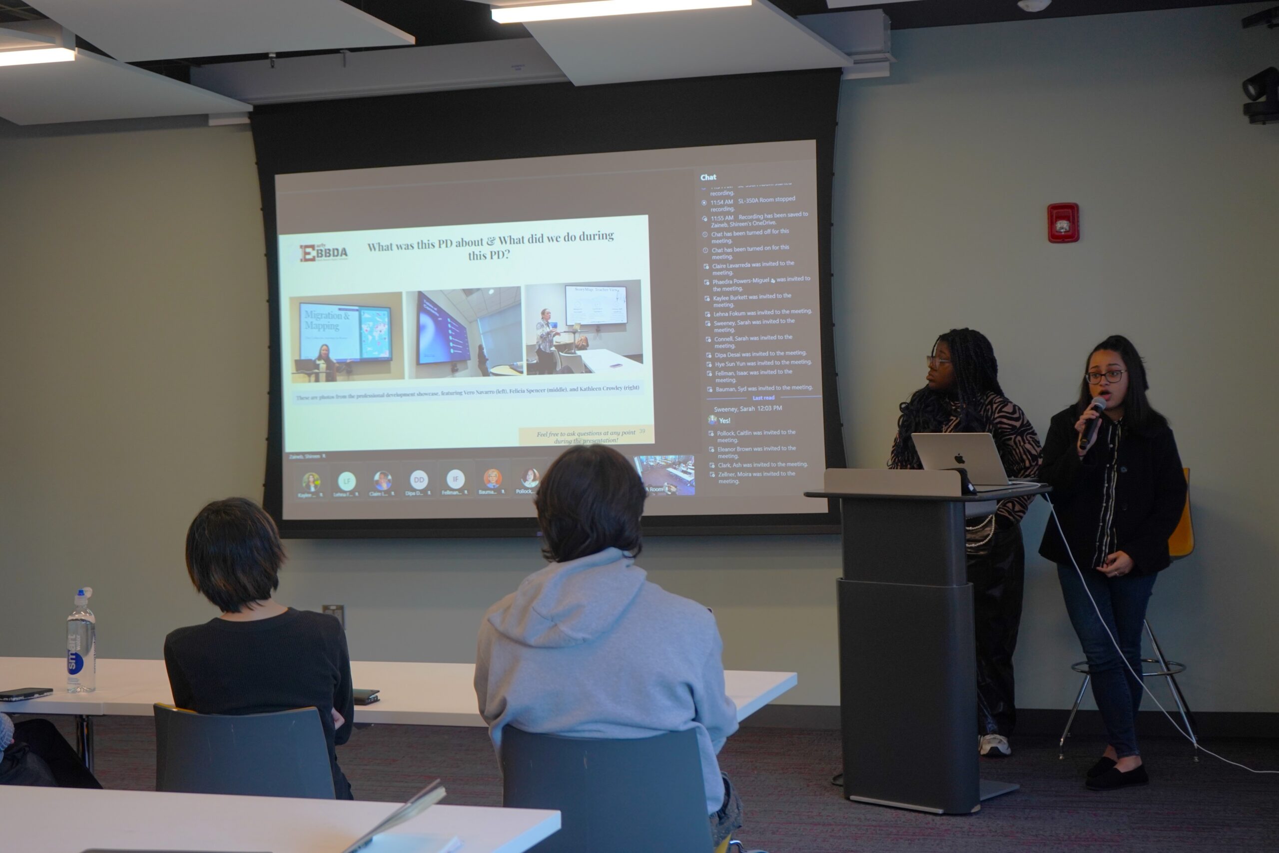 Savita discussing the professional development workshop. Two individuals standing behind a podium, with the person on the right holding the microphone. There are projected slides behind them and two audience members visible from behind.