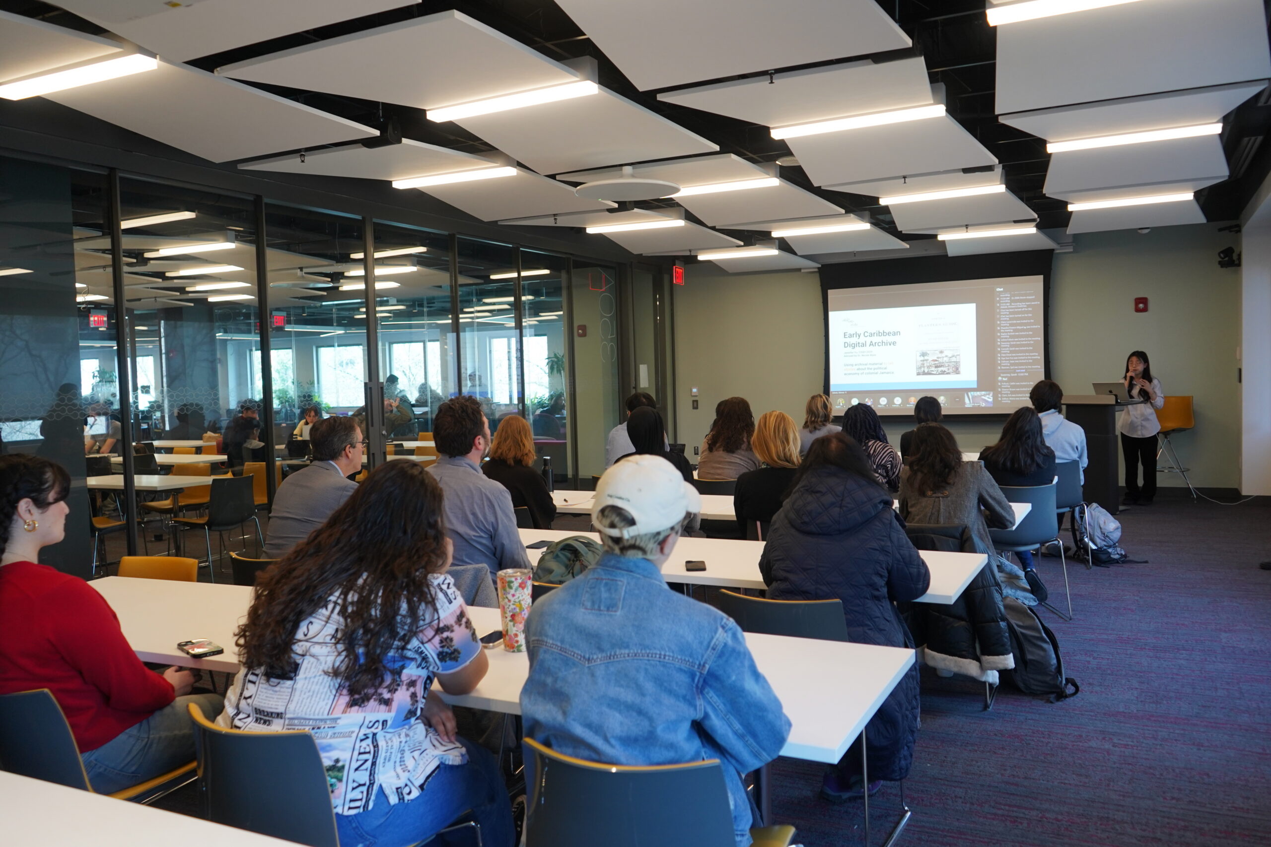 An individual standing behind a podium on the left holding a microphone. There are slides displayed behind. There are seventeen audience members visible from behind.